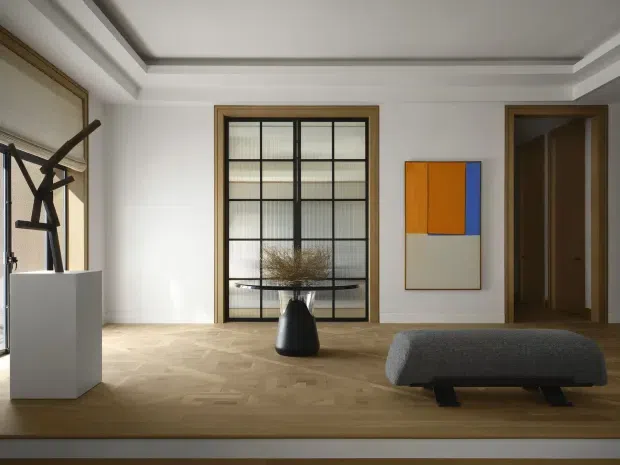 A refined contemporary interior, likely a living room or gallery-like sitting area, with a pale herringbone timber floor, white walls, a detailed recessed tray ceiling, black-framed gridded internal glazing, and warm timber door and window surrounds. Minimal furnishings include a low dark upholstered bench, a sculptural black vase with dried branches, a white plinth supporting abstract artwork or sculpture, and a colourful geometric wall artwork. The room is bright, restrained, and art-focused.