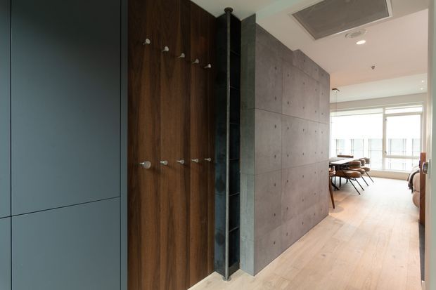 Interior view of a modern residential hallway or entry area, featuring a dark teal built-in cabinet wall, warm timber coat-storage panel with hooks, a narrow dark tiled shower-like or storage recess, and a large exposed concrete partition wall. Pale timber flooring leads toward a bright dining or living area with expansive glazing. The space has a clean, minimalist contemporary fit-out with contrasting raw and warm materials.