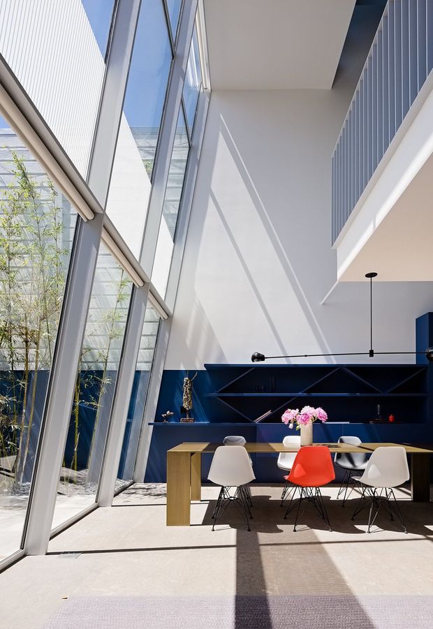 Bright contemporary double-height dining area with floor-to-ceiling angled glazing, a deep navy built-in shelving wall, a long timber dining table, and modern molded dining chairs including a vivid red accent chair. Strong sunlight creates geometric shadows across the pale floor, while an interior planted courtyard is visible beyond the glazing.