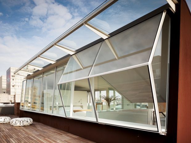 Exterior view of a contemporary glazed rooftop terrace or sunroom enclosure, featuring extensive angled glass panels in dark metal frames, operable awning-style glazing, a timber deck, and dark brown wall cladding. Modern apartment or commercial buildings are visible in the background.