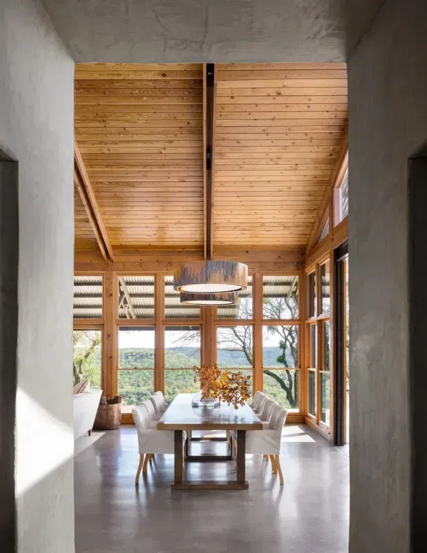 A refined dining area viewed through a concrete-framed opening, featuring a long timber dining table with upholstered chairs, expansive timber-framed glazing, a pitched exposed timber-lined ceiling, and two circular pendant lights. The room opens to views of a wooded, hilly landscape and receives abundant natural light. Concrete walls and floor contrast with the warm timber structure and joinery.