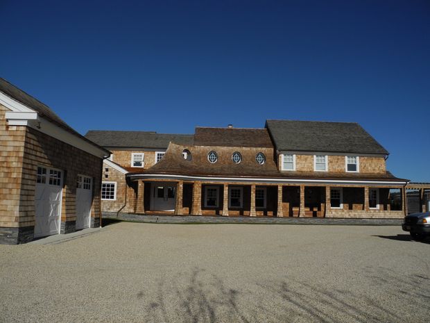 Exterior view of a large traditional shingle-clad building arranged around a broad gravel courtyard. The main two-storey structure has steeply pitched dark roofs, dormer-like round windows, white-trimmed sash windows, and a long covered veranda with multiple entry doors. A matching outbuilding with garage doors is visible at left, while a partial vehicle appears at the right edge.