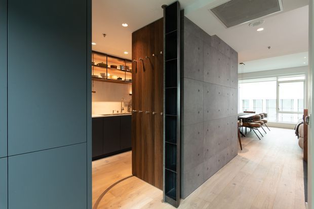 A contemporary open-plan interior viewed from a circulation area, featuring a dark teal built-in wall at left, a warm timber pivot or storage panel with open shelving, a compact kitchen beyond, and a raw concrete feature wall dividing the kitchen from a dining or living area. Pale timber flooring, recessed lighting, and broad glazing create a refined industrial-modern atmosphere.