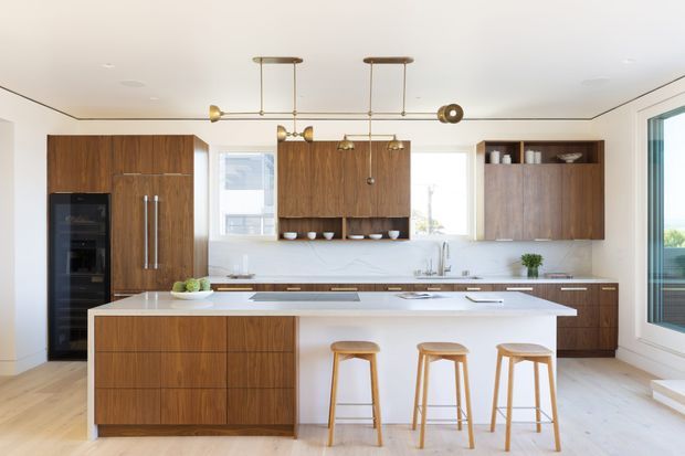 Bright contemporary kitchen with extensive warm timber cabinetry, a large white island with three timber stools, integrated appliances, white benchtops, a subtle white splashback, and brass-toned linear pendant lighting. Large windows and clean detailing create an airy, refined interior.