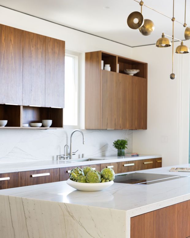 Bright contemporary kitchen featuring warm walnut-toned flat-panel cabinetry, a large white veined stone island and backsplash, integrated cooktop, and a sculptural brass and bronze multi-light pendant. Open shelving displays simple ceramics, while a large window adds natural light.