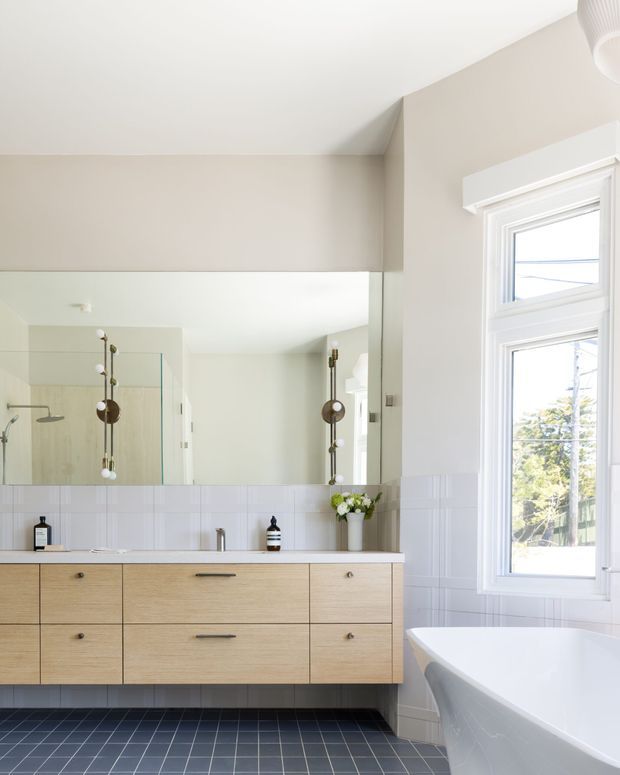 Bright, refined bathroom with a large white-framed window, freestanding white bathtub, wall-to-wall mirror, glass shower enclosure, and a long floating timber vanity with integrated white basin. Pale warm-grey walls and white tiled wainscoting contrast with the dark blue-grey tiled floor. Minimal botanical and apothecary-style accents add a calm, contemporary feel.