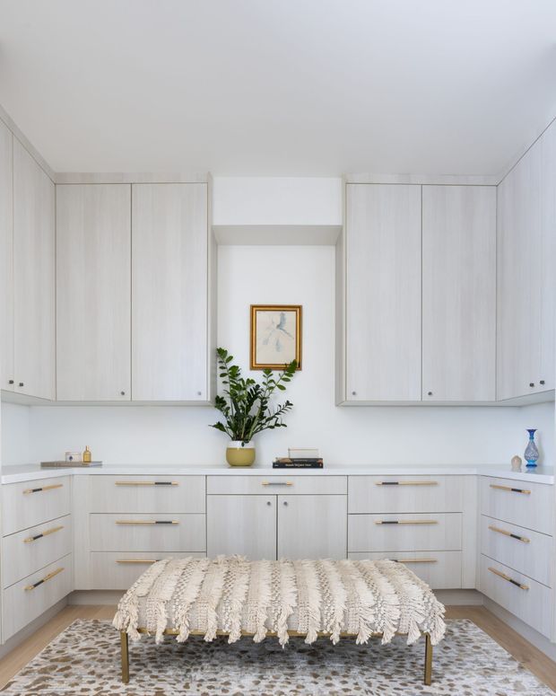 Bright, minimalist dressing or bedroom storage area with extensive pale wood built-in cabinetry wrapping three walls. A central white wall niche displays framed artwork and a potted plant, while a low upholstered bench with a textured cream throw sits on a patterned rug in the foreground. The space has a clean, restrained palette with brass-toned cabinet pulls and abundant integrated storage.