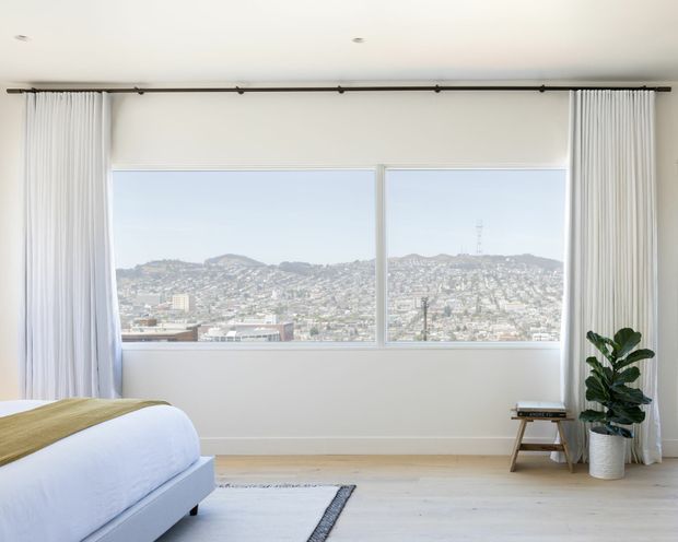 Bright, minimalist bedroom with a low upholstered bed, pale timber flooring, white walls, full-height white curtains on a dark track, and a large picture window overlooking a densely built hillside urban landscape. A potted plant and small wooden stool add understated natural accents.