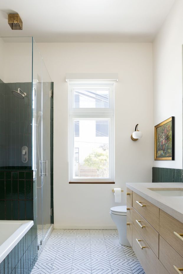 A bright, narrow bathroom featuring a walk-in glass shower with dark green vertical wall tiles, a white bathtub, patterned geometric floor tiles, a timber vanity with a pale stone benchtop, a toilet, and a tall white-framed window. Brass-toned hardware, a wall sconce, and framed artwork add warm decorative accents.