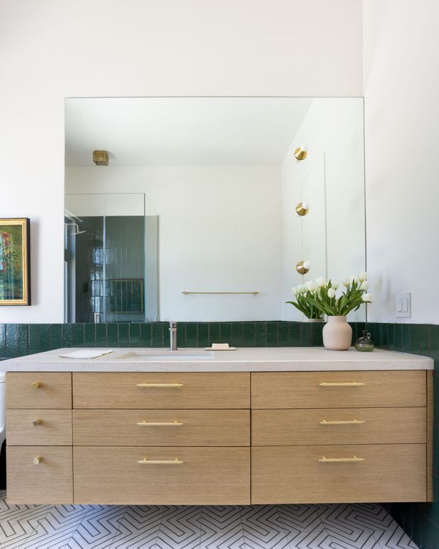 A stylish bathroom vanity wall with a large frameless mirror, wall-mounted light timber drawer cabinetry, a pale stone benchtop, integrated basin and brass tapware. Deep green rectangular wall tiles form the backsplash and continue around the room, while patterned geometric floor tiles add visual interest. Brass wall sconces, a framed artwork, and a vase of tulips provide decorative accents.