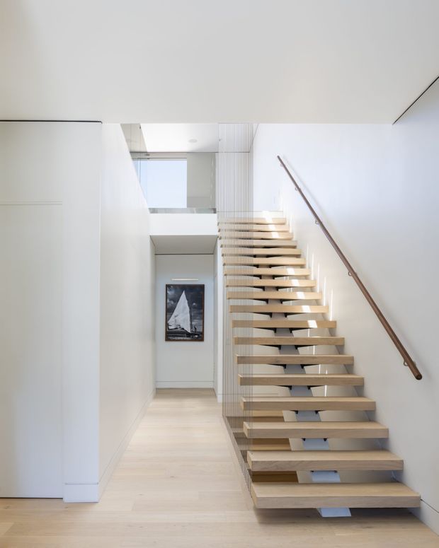 A bright minimalist interior hallway focused on a floating open-riser staircase. Pale timber treads extend along a white wall with a slim timber handrail, while a vertical slatted screen forms the stair balustrade. Light timber flooring, white walls, and a framed artwork create a clean, airy composition. An upper-level opening and glazing bring additional daylight into the stairwell.