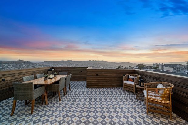 A rooftop terrace overlooking a densely built hilly urban landscape at sunset. The terrace features patterned geometric floor tiles, a dark timber privacy balustrade, a dining table with woven chairs, and a small wicker lounge seating area. The scene has expansive sky views and a warm, inviting outdoor entertaining atmosphere.