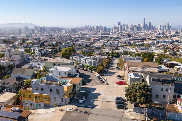 Aerial view over a dense urban residential neighbourhood, with rows of low-rise homes and apartment buildings in the foreground and a prominent high-rise city skyline in the distance. Streets, rooftops, parked vehicles, and varied modern residential architecture are clearly visible under bright daylight.