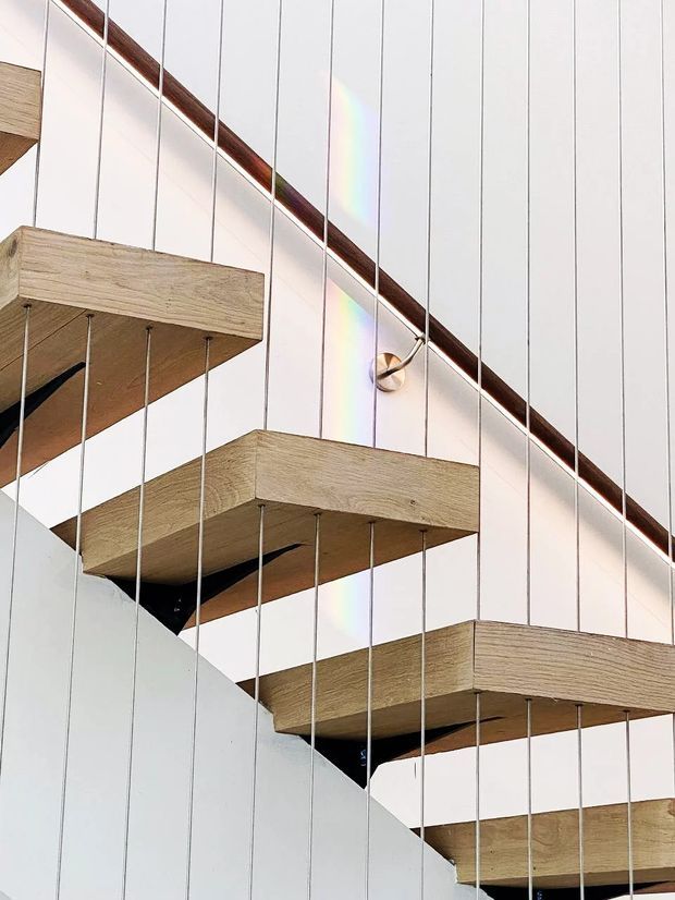 Close-up architectural view of a modern suspended staircase. Thick light timber stair treads appear cantilevered from a white wall, with numerous slender vertical tension rods supporting or stabilising the treads. Dark recessed structural elements are visible beneath the steps, along with a curved timber handrail or trim and a small wall light. The composition is bright, minimal, and highly geometric.