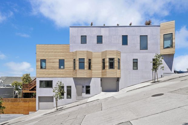 Exterior view of a modern multi-storey residential building on a steeply sloping urban street. The facade combines pale grey smooth wall panels with horizontal light timber cladding, projecting window bays, dark-framed rectangular windows, and an integrated lower-level garage. A neighboring timber fence and houses are visible at the sides under a bright blue sky.