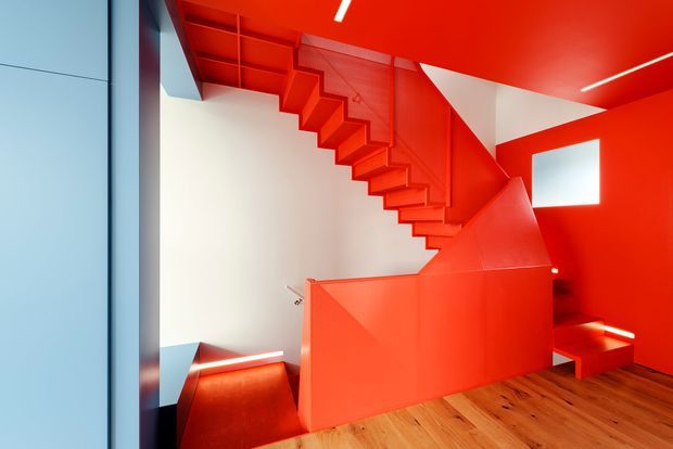 A striking contemporary stairway interior dominated by vivid red painted surfaces. A sculptural open-riser staircase runs diagonally across the frame, with angular red balustrades and enclosing walls. Warm timber floorboards contrast with the red ceiling, walls, and stair structure, while integrated linear lighting and a small square window add visual accents. The space appears compact but vertically dramatic, with strong geometric forms and high contrast.