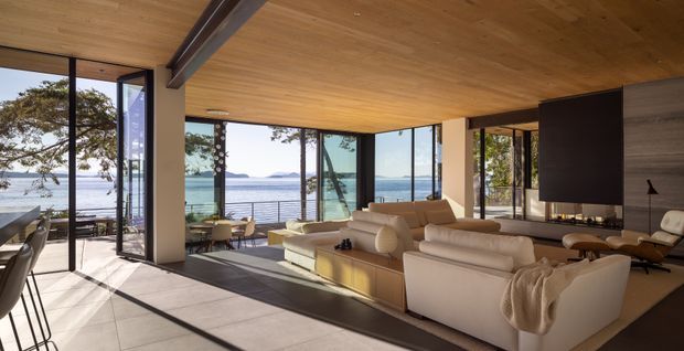 A spacious, open-plan living room with floor-to-ceiling glazing overlooking a calm waterfront. The room features a warm timber-lined ceiling, pale tiled flooring, dark structural beams and window frames, a large cream sectional sofa, a dark built-in media wall, and a dining area extending toward the glazed balcony or terrace. Strong indoor-outdoor connection and abundant natural light define the space.