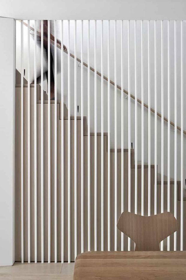 A minimalist interior view of a staircase screened by closely spaced vertical white slats. Pale timber stair treads and a timber handrail are visible behind the screen, with a sculptural timber dining chair and rounded timber tabletop in the foreground. The composition is clean, restrained, and softly lit, with strong linear rhythm and neutral materials.
