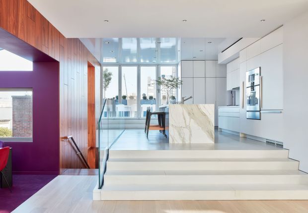 Bright contemporary open-plan interior with a stepped transition from a lower living area into a raised kitchen and dining zone. The space features extensive white built-in cabinetry, a marble-look waterfall island, pale timber flooring, dark reddish timber wall panelling, a deep purple accent wall and large glazed openings at the rear. Recessed ceiling lighting and clean linear detailing create a refined, minimalist appearance.