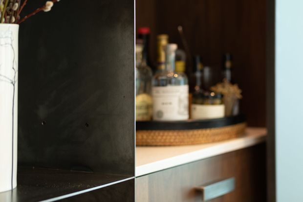 Close-up of a dark, refined built-in bar or drinks cabinet. Bottles and glassware sit on a round tray within open shelving, with a reflective dark feature panel at left, a pale horizontal shelf edge, and a wood-toned cabinet front with a metal handle below. The image is tightly framed and softly lit, emphasizing the joinery and bar contents.