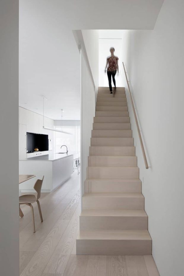 Bright minimalist interior view focused on a straight staircase with pale timber treads and matching pale timber flooring. A simple timber handrail runs along the white wall, with a white balustrade on the opposite side. The staircase rises to an upper landing, while an open-plan white kitchen and part of a dining area are visible beside it. The image has clean lines, white surfaces, light wood, and abundant natural light.