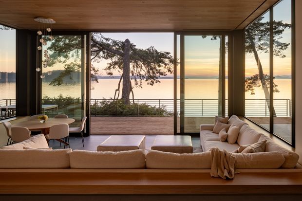 A refined open-plan living and dining room with a warm timber ceiling and extensive floor-to-ceiling glazing overlooking calm coastal water at sunset. A large cream sectional sofa faces the view, with a low timber platform or built-in base, while a compact dining table and upholstered chairs sit to the left. Slim dark window and door frames, a timber deck, and simple horizontal balcony railings reinforce the strong indoor-outdoor connection.