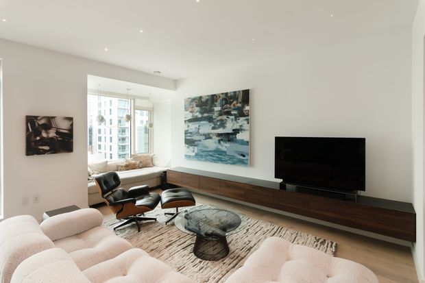 Bright contemporary living room with a low-profile modular cream sofa, iconic lounge chair and ottoman, textured neutral area rug, glass coffee table, and long dark wood wall-mounted media console with television. A large abstract artwork and a window seat by expansive glazing add visual interest. The room has recessed ceiling lighting, pale walls, and an urban apartment outlook.