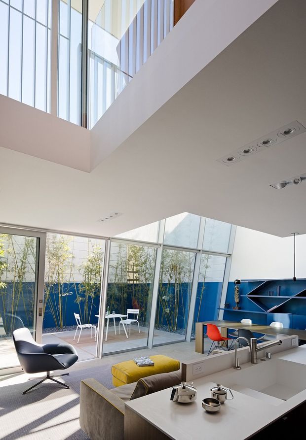 Bright, double-height contemporary open-plan living space with a white kitchen island in the foreground, a lounge area with a dark lounge chair and sectional seating, and a dining/work table beyond. Floor-to-ceiling glazing opens onto a small courtyard, while a bold blue built-in shelving wall and colourful seating provide accents. Recessed ceiling lighting and a large overhead void emphasize the airy architectural volume.