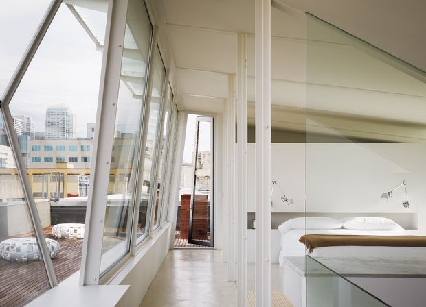 Bright contemporary apartment interior with a glazed bedroom separated from a narrow circulation space by full-height glass and white-framed partitions. The bedroom contains a low platform bed with white bedding and a slim wall-mounted reading light. Angled floor-to-ceiling windows open toward a small timber deck and an urban cityscape, while polished pale flooring, white walls, and extensive glazing create an airy, minimalist feel.