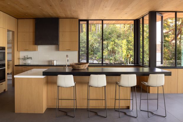 A warm, open-plan contemporary kitchen with extensive light timber cabinetry, a large island with dark benchtop and four bar stools, and a dark tiled floor. Full-height black-framed windows and glazed doors overlook leafy vegetation, while timber continues across the ceiling. The kitchen includes integrated appliances, a black rangehood, undermount sink, and a generous breakfast bar. The space is bright, refined, and strongly connected to the garden setting.