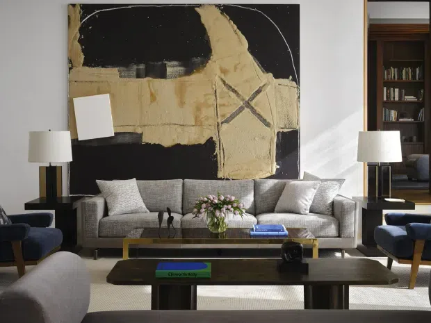A refined living room with a large abstract black, cream, and metallic artwork above a grey upholstered sofa. Two matching table lamps flank the seating, while layered wood and dark coffee tables, blue accent armchairs, cushions, and a floral arrangement create a balanced, contemporary interior. A doorway in the background reveals dark timber joinery and shelving in an adjoining space.