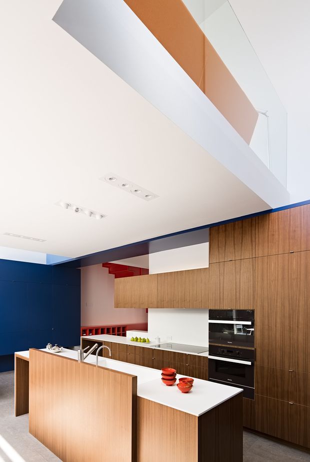 A bold contemporary kitchen with a large white island, integrated sink and tap, warm vertical-grain timber cabinetry, and built-in black appliances. Deep blue wall panels and red accents add strong colour contrast. The space has a bright white ceiling with recessed linear spotlights and a high open void or upper-level opening above the cabinetry, creating a crisp, architectural interior.