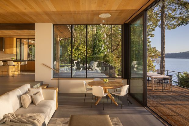 A warm, contemporary open-plan living and dining area with extensive floor-to-ceiling glazing opening onto a timber deck. The room features a timber-lined ceiling, neutral upholstered seating, a round timber dining table with modern chairs, dark flooring, and a sculptural pendant light. Through the glazing, a wooded waterfront setting and lake are visible.