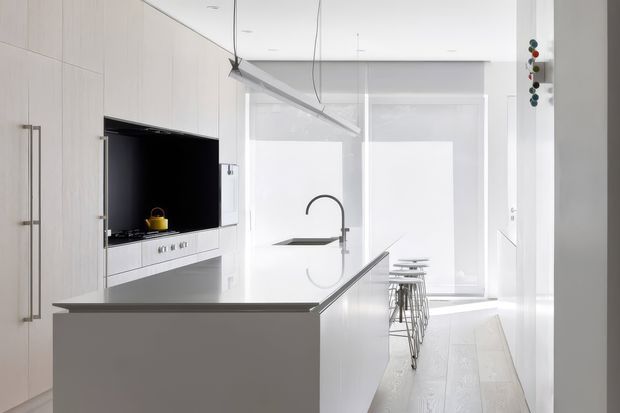 Bright, minimalist contemporary kitchen with extensive pale built-in cabinetry, a large island with integrated sink and sculptural black faucet, and a wall-mounted television. A linear suspended light runs above the island, while bar stools sit at a compact dining or breakfast counter near full-height windows with roller blinds. The palette is predominantly white and light grey with black accents and abundant natural light.