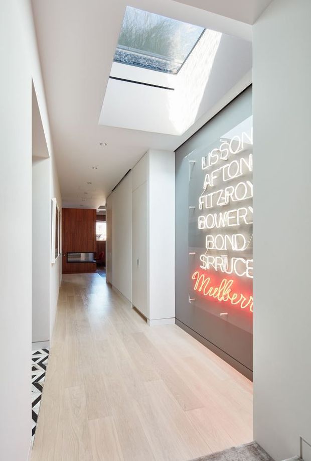 A bright, minimalist residential hallway with pale timber flooring, smooth white walls and concealed cabinetry. A large skylight brings daylight into the corridor, while a full-height dark glass or panelled feature wall displays illuminated white and red neon lettering. A warm timber-lined room is visible at the far end, and a patterned floor edge appears through an opening on the left.