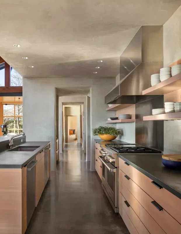 A narrow, elongated contemporary kitchen viewed down a central passage. The room features warm timber cabinetry with dark linear handles, dark countertops, a stainless-steel range and large sculptural stainless-steel range hood, open timber shelves displaying white crockery, a concrete-look ceiling and walls, polished dark flooring, and timber-framed glazing at the rear. Recessed downlights and natural light create a refined, warm industrial atmosphere.