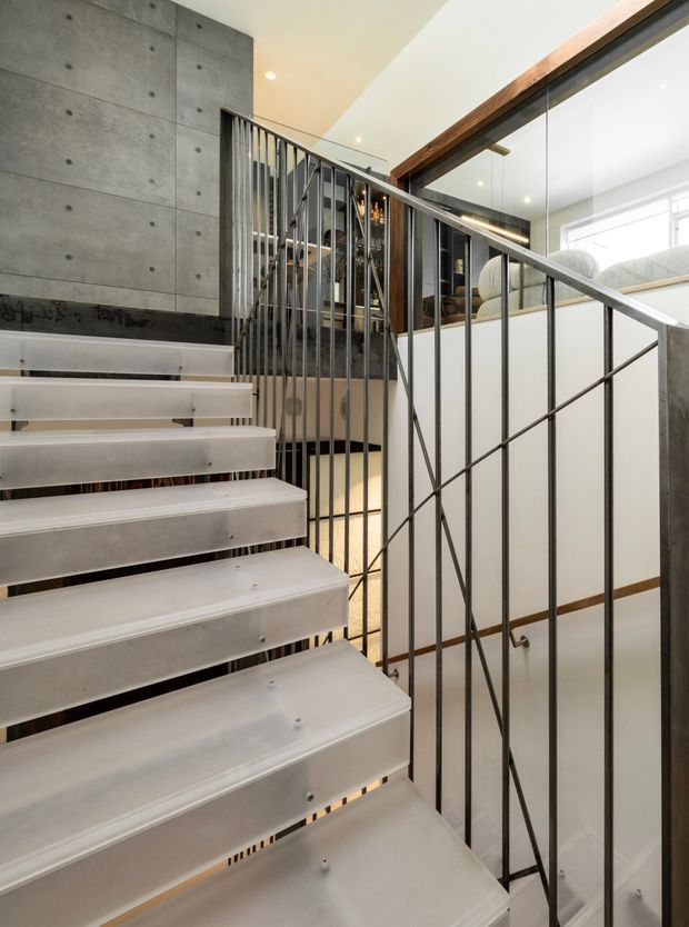 Interior view looking up a contemporary stairway with open-riser pale steps, a dark metal balustrade with diagonal bracing, and a polished concrete feature wall. A glass-partitioned upper-level space and warm timber trim are visible beyond the stairs.