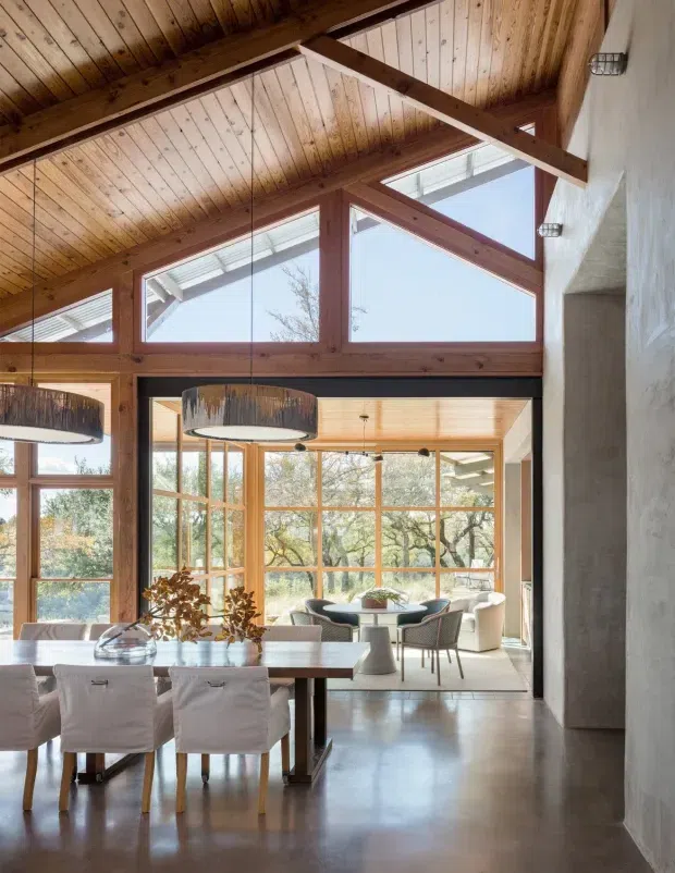 A bright open-plan dining and living space with a large timber-framed glazed wall, exposed pitched timber ceiling and structural beams, polished concrete flooring, and a dining table in the foreground. The room opens toward a light-filled sitting area surrounded by windows, with views of trees outside. Contemporary furnishings are paired with warm natural timber and plaster walls.