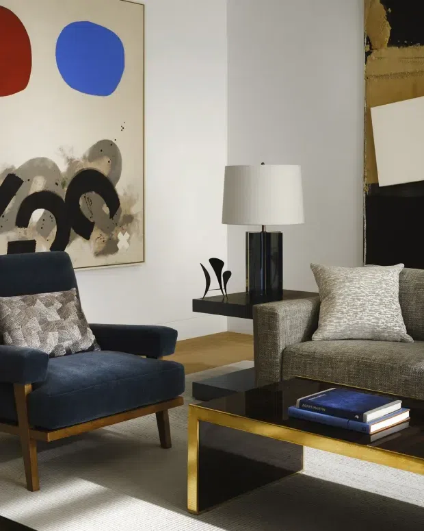 A refined living room featuring a deep navy upholstered lounge chair, a textured taupe-grey armchair, and a rectangular coffee table with a dark glass top and brass-coloured metal frame. A sculptural black side shelf supports a white drum-shade table lamp, while an abstract artwork adds red, blue, black, and neutral tones to the pale wall. The room has a warm, curated mid-century-inspired aesthetic with strong geometric forms.