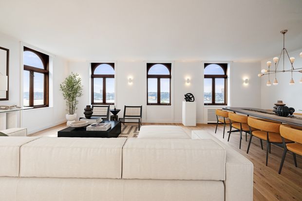 Bright, spacious open-plan apartment interior combining a living area and dining zone. The room has white walls and ceiling, pale timber flooring, a large cream sectional sofa, black coffee table, sculptural occasional chairs, and a long dining table with mustard/tan upholstered chairs. Tall arched-top windows with dark timber frames overlook a waterfront or urban skyline, while a contemporary branching pendant light and minimal wall sconces provide lighting. The overall aesthetic is refined and contemporary with strong mid-century-inspired furniture accents.