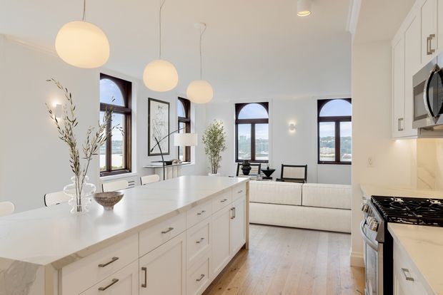 Bright open-plan interior combining a white kitchen with a large central island and an adjoining living area. The room has white shaker-style cabinetry, pale stone benchtops, warm timber flooring, tall dark-framed arched-top windows, and globe pendant lights. A light sofa and simple contemporary furnishings sit beyond the island, with views visible through the windows.
