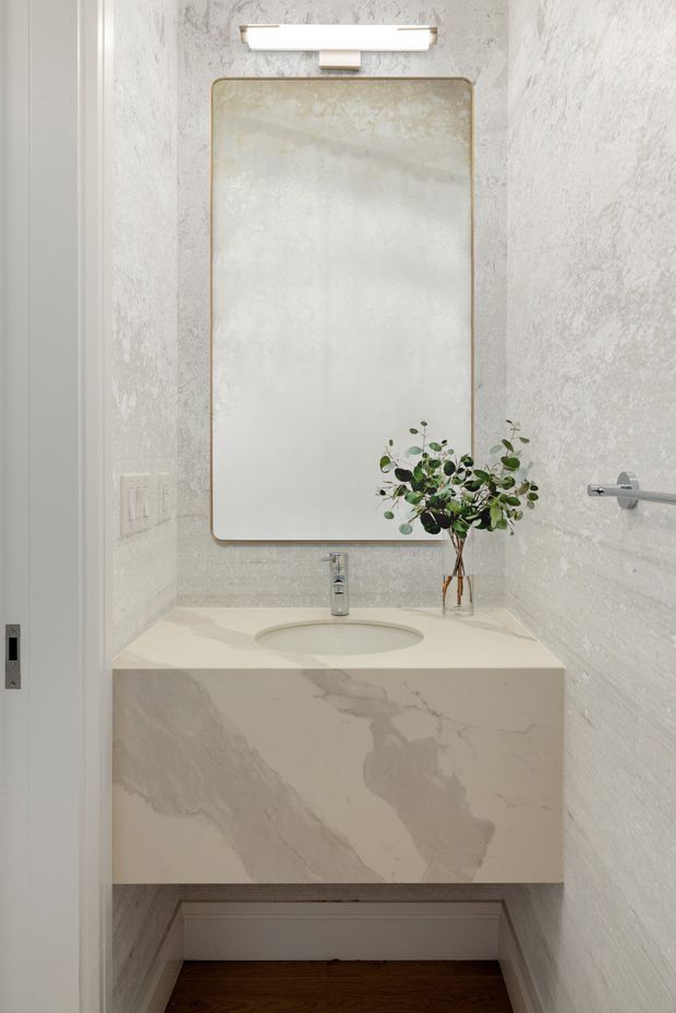 A compact, refined bathroom vanity area with a floating stone vanity and integrated oval basin. The walls are covered in textured pale stone-like finish, complemented by a large rounded-rectangle mirror with a slim brass-toned frame and a contemporary horizontal wall light. A chrome tap and towel rail add functional fixtures, while a small vase of greenery provides a natural accent. Dark timber flooring is visible beneath the vanity.