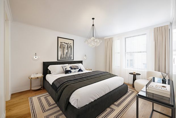 Bright, refined bedroom with a centered upholstered bed, dark charcoal bedding and headboard, layered geometric area rug, white walls with decorative ceiling and wall trims, and tall bay-style windows dressed with sheer curtains and beige drapes. A sculptural clustered pendant hangs above the bed, with slim bedside tables, a small upholstered chair, and a compact desk or console completing the room.