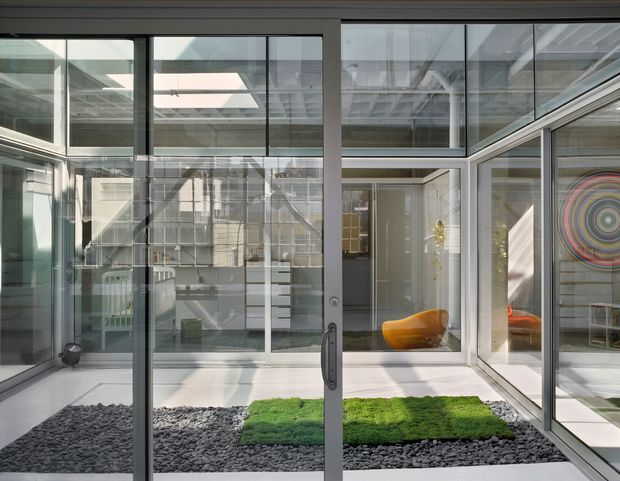 Bright modern glazed courtyard or atrium viewed from inside, with extensive black-framed floor-to-ceiling glass, a white structural frame, skylight and visible exposed services. The central indoor-outdoor space contains dark pebbles and vivid green planted patches, while adjoining contemporary interiors and colourful art are visible through the glazing.