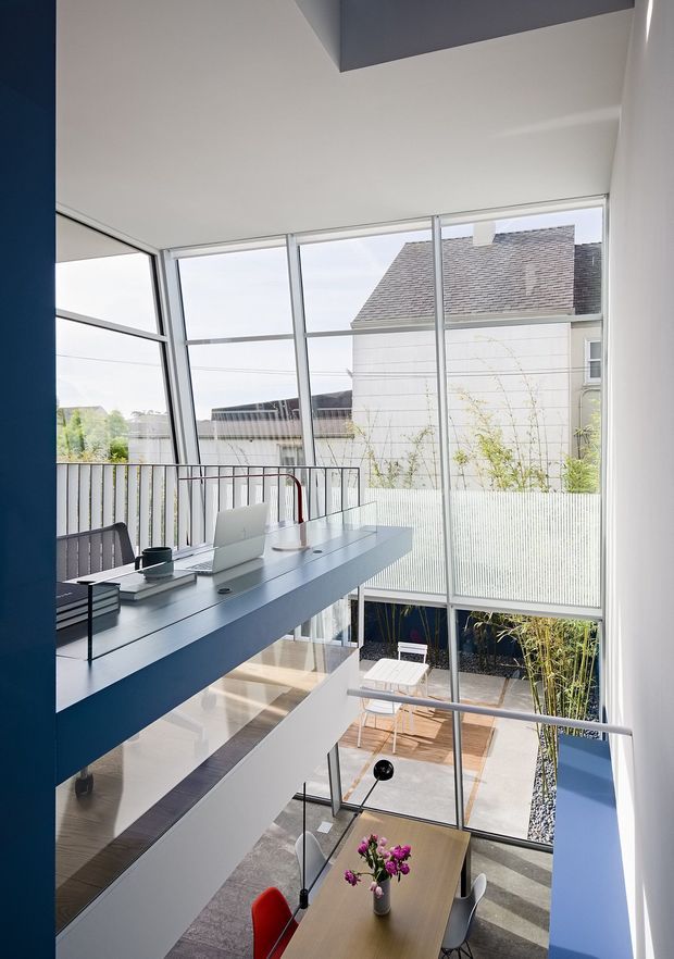 A bright, multi-level contemporary home office overlooking a small courtyard through expansive floor-to-ceiling glazing. A long blue built-in desk with a laptop and task items projects over the lower dining area, while a stair or landing with slim metal balustrades frames the view. The interior has white walls and ceilings, blue accents, timber flooring and decking, and a compact outdoor seating area visible below.