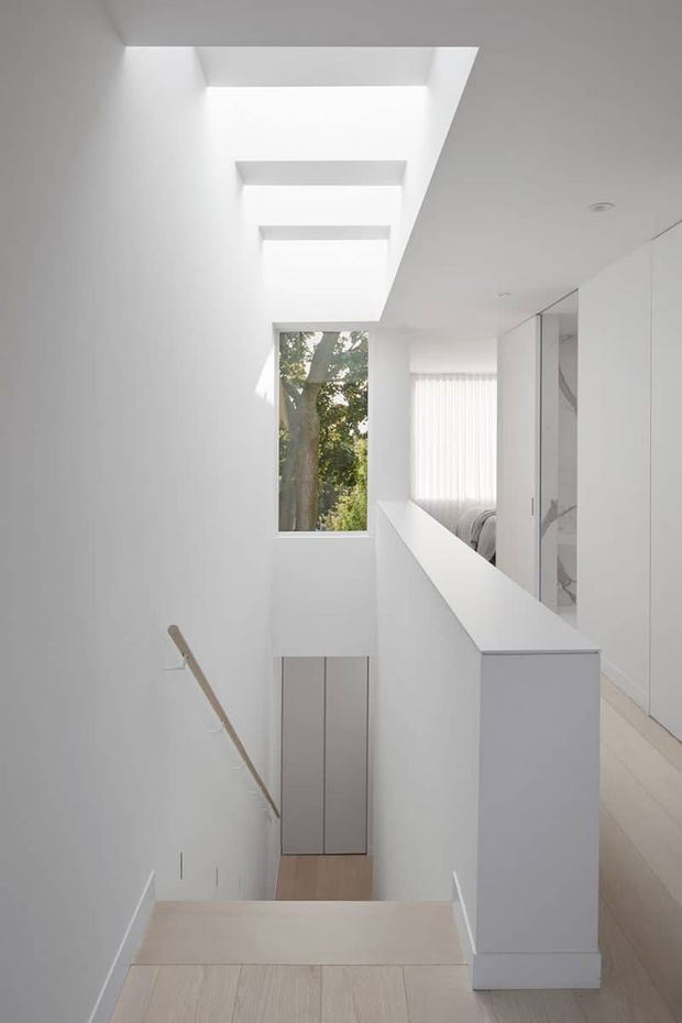 Bright minimalist interior view from the top of a stairway, looking down toward a lower landing. The space has crisp white walls and built-in cabinetry, pale timber stair and floor finishes, a simple timber handrail, a high vertical window, and multiple overhead skylights that flood the stairwell with natural light. A bedroom area is partially visible beyond the half-height partition, with white cabinetry and a marble-look vertical surface.