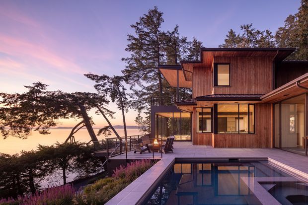 A modern multi-level timber-clad residence set on a sloping waterfront site, photographed at sunset. The home features strong horizontal rooflines, expansive glazing, a cantilevered upper volume, an elevated terrace with outdoor seating, and a rectangular swimming pool in the foreground. Mature trees and the calm water beyond create a secluded coastal setting.