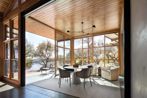 A bright, glazed sunroom or enclosed terrace with extensive timber-framed windows overlooking a landscaped waterfront setting. The room has a timber-lined ceiling, pale tiled floor, large rug, round dining table, woven dining chairs, lounge seating, and a contemporary black multi-arm pendant light. Open glazing connects the interior to an outdoor terrace, with strong natural light and views of water and trees.