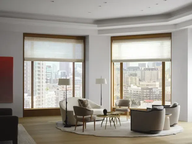 A refined high-rise living room with expansive city views through two large timber-framed windows. The room features pale timber flooring, white walls, layered recessed ceiling detailing, a neutral upholstered seating arrangement, sculptural floor lamps, small round coffee tables, and a large organic-shaped rug. The interior has a quiet, sophisticated contemporary character with strong natural daylight.