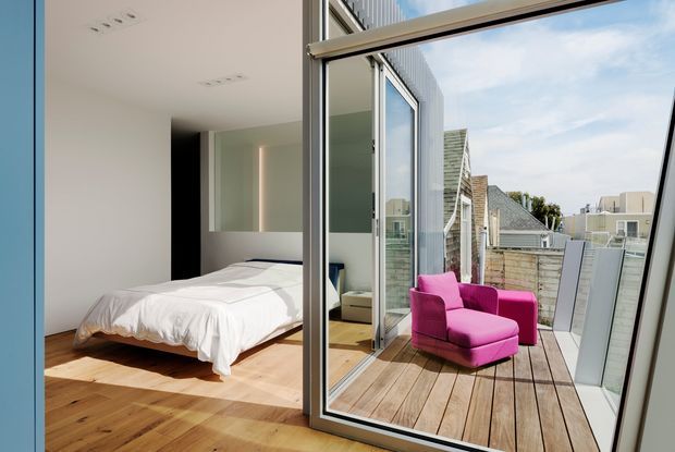 A bright modern bedroom opens directly onto a timber rooftop terrace through large glazed doors. The room has a simple white bed, pale walls, dark vertical feature panel, and warm timber flooring, while the terrace includes a vivid pink lounge chair and views of neighbouring urban buildings. The composition strongly emphasizes indoor-outdoor connection and extensive glazing.
